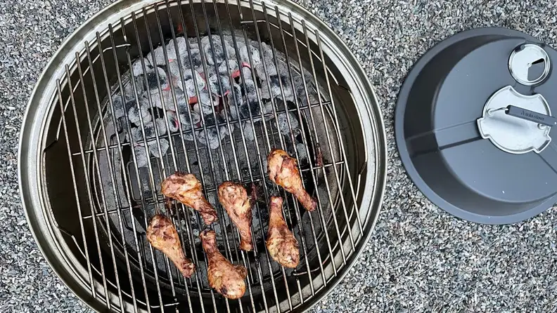 Chicken drumsticks cook on a grate over a charcoal grill.