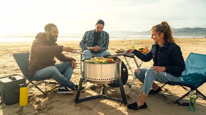 Three people grill vegetables on a Solo Stove while camping on a beach.