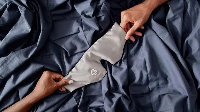 Weighted sleep mask being stretched out over bed by two hands.