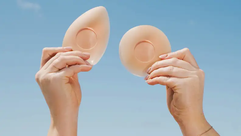 Hands holding two different shapes of Cakes nipple covers against a clear blue sky.