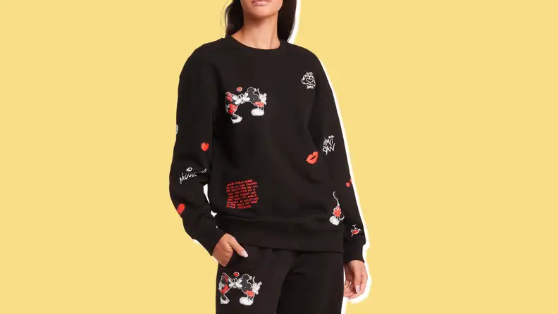 A woman wearing a black crewneck sweatshirt with a picture of Mickey and Minnie Mouse kissing and red and white writing against a yellow background.