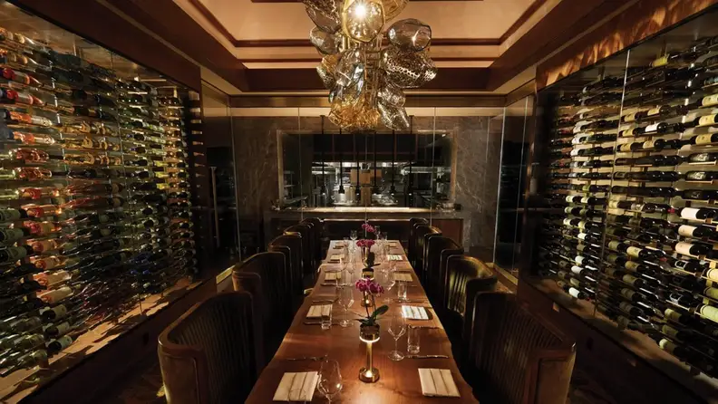 Long dining table with elegant table setting, racks of wines on the left and right wall, plus wall-to-wall mirror, all in a dimly-lit private room