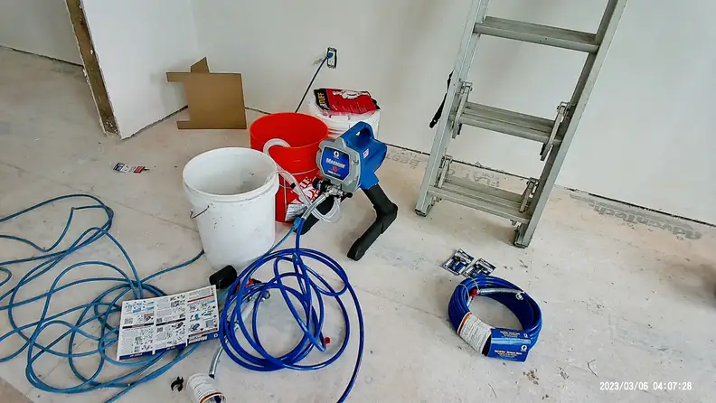 A Graco Magnum X5 Airless Paint Sprayer next to an orange and white bucket in a room under construction.