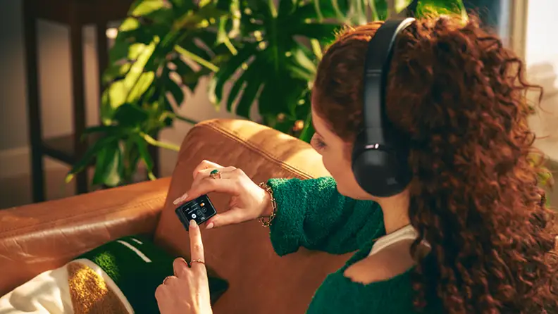 Woman sitting on a couch listening to JBL Tour One headphones controlling the music with included handheld control device.