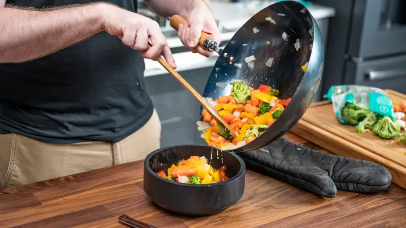 A person is pouring out a medley of vegetables from a wok into a bowl.