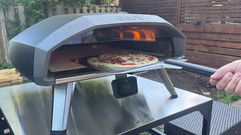 An Ooni outdoor pizza oven sits on a stainless steel table with a pepperoni pizza