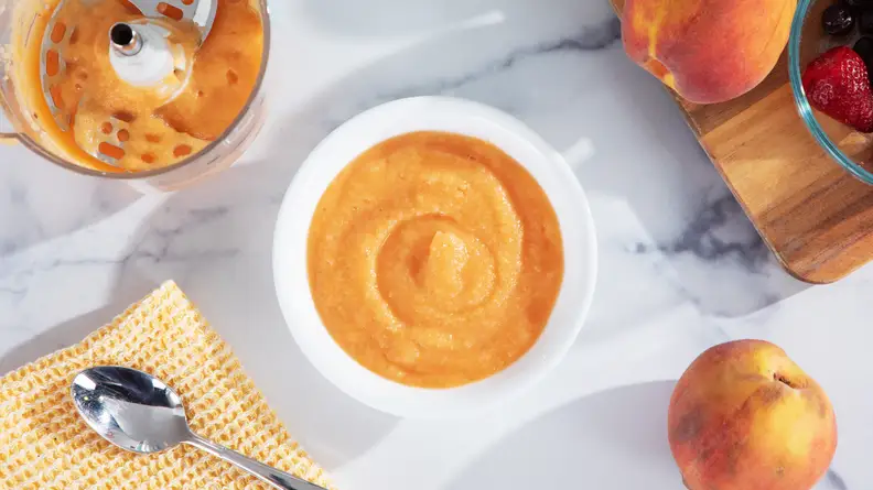 A bowl of puree peaches next to the Nutribullet Baby Steam + Blend containers and peaches on the side.