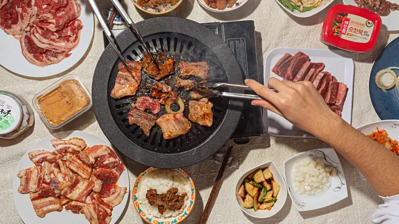 A typical Korean barbecue spread with a iron barbecue stove in the middle, grilling sliced pork belly. Around the stove, there are plates of thinly sliced meat, pickled vegetables, and sauces.