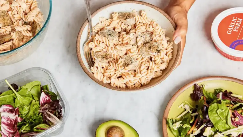 Bowl of creamy garlic parmesan chicken pasta served alongside mixed greens and fresh ingredients on a countertop.