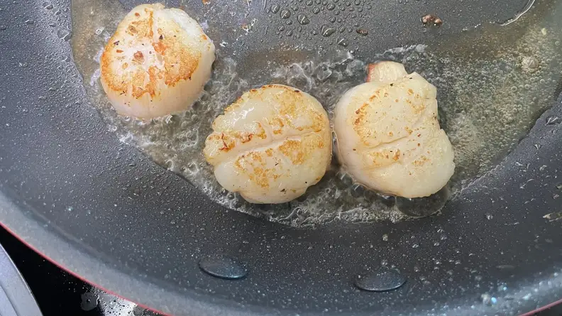 Three small scallops searing inside of greasy skillet pan.