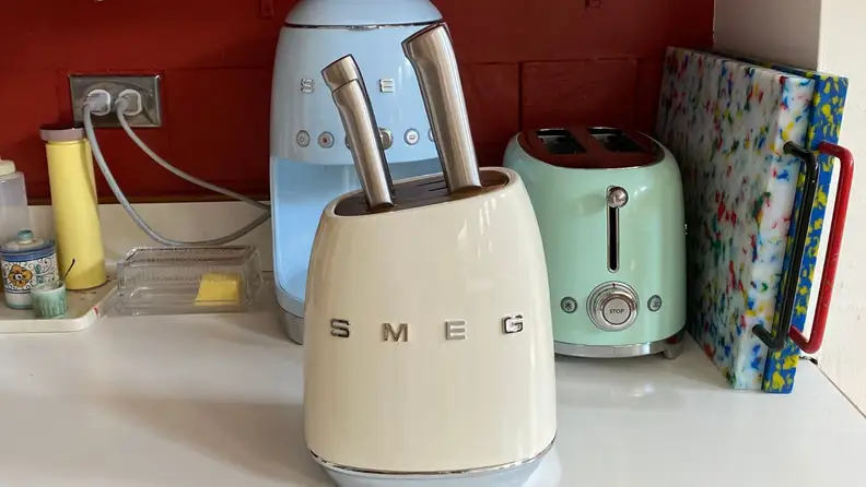 A cream colored Smeg knife block set sitting on a kitchen counter in front of other Smeg appliances.