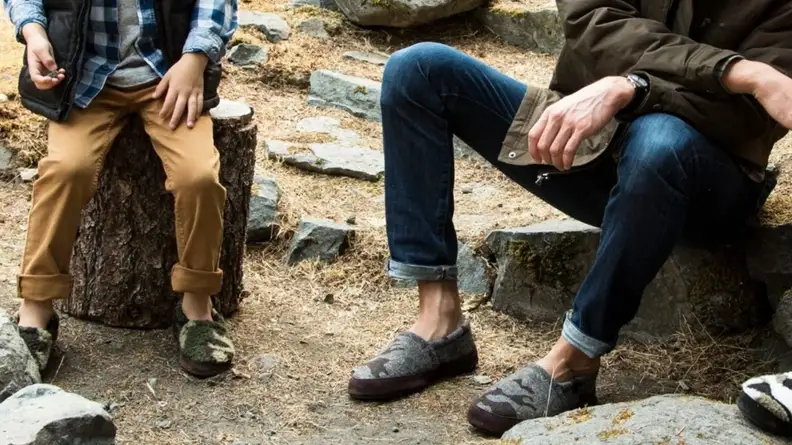 A child and a man sitting together, each wearing a pair of Acorn Moc Slippers in men's sizes.