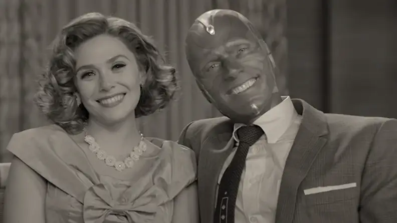 A black and white still from WandaVision featuring Wanda Maximoff and Vision on a sofa in 50s clothes.