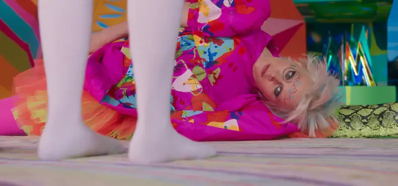 Weird Barbie, played by Kate McKinnon, lays sideways on the ground at Stereotypical Barbie (Margot Robbie's)'s feet