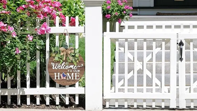 A wooden "Welcome to our home" sign hangs on a white fence post, featuring a patriotic star as the "O" in "home"