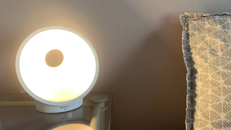 Image of the Philips Connected sunrise alarm at full brightness in a room on a clear end table next a bed with a grey pillow