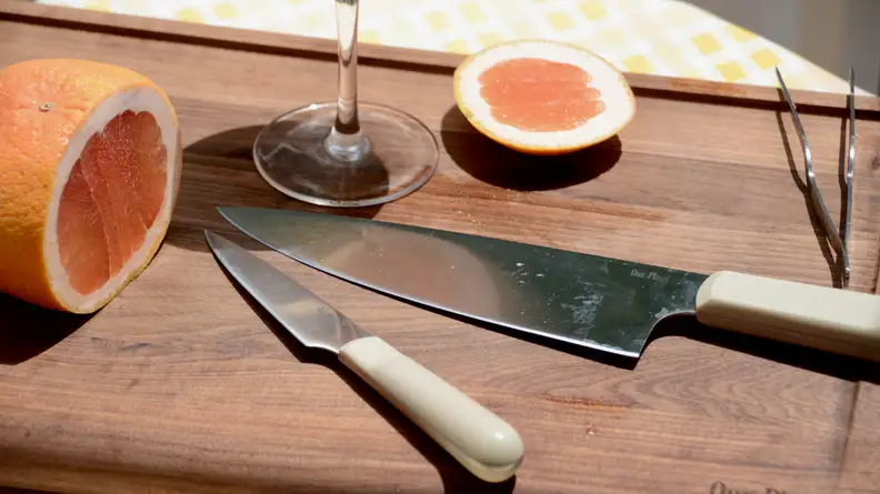 On an Our Place walnut wood cutting board, there's a half-sliced grapefruit and a wine glass. Next to these items, there's a paring knife and a chef's knife by the same brand.