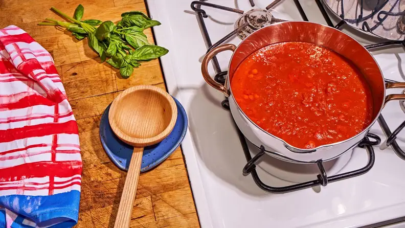 A pot of tomato sauce simmers on a gas cooktop with a wooden ladle to the left perched on a blue spoon rest.