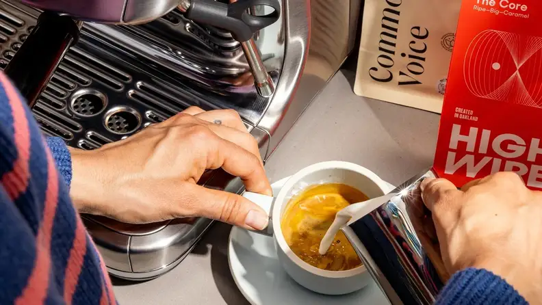 Hands pouring milk into a latte beside Common Voice and High Wire coffee bags