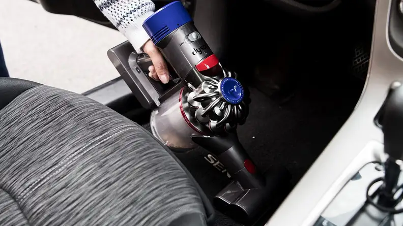 A close up shot of a person using the Dyson V8 vacuum to clean the floor of a motor vehicle.