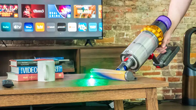 A close up of a person using the Dyson V15 vacuum to clean food off a coffee table. There is a TV in the background.