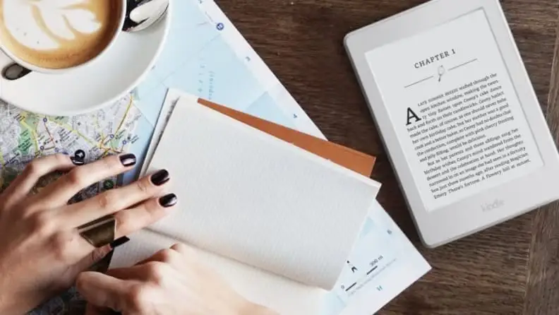 A woman's hand with a dark manicure holds a notebook open beside a coffee cup and beside a white Kindle Paperwhite e-reader