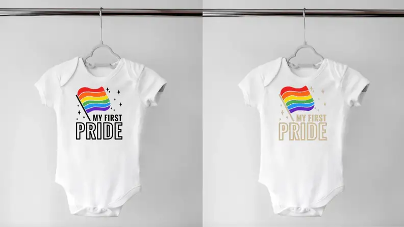 Two white onesies hanging on a hanger with a rainbow flag on it and the words "My First Pride" written on it