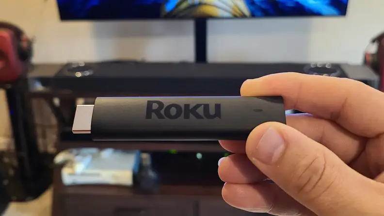 The matte black, candy-bar sized Roku Streaming stick is held with its port showing an OLED TV.