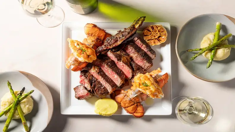 Choice steak salted and cut into slices, and lobster tails on a square plate, two plates of mashed potatotes with asparagus, and two glasses and a bottle of white wine on a clothed table