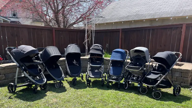 Seven different strollers lined up in a backyard.