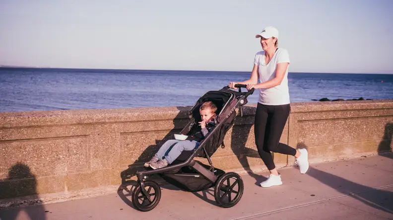 A woman jogging along a sea wall pushing a child in a jogging stroller.