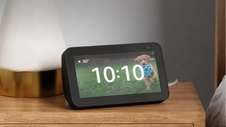 The third-gen Amazon Echo Show 5 with the time displayed on the screen, sitting on a wood nightstand.