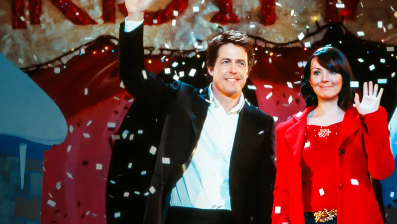 A bashful Prime Minister (Hugh Grant) and aide-turned-love interest Natalie (Martine McCutcheon) wave at a school auditorium crowd after being caught kissing back stage.