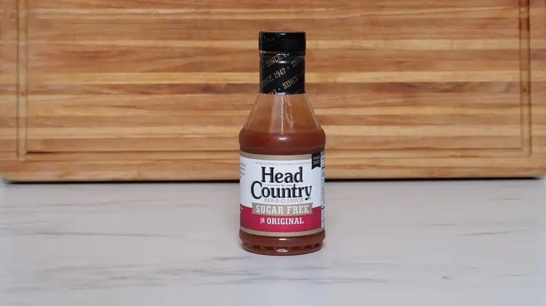 A bottle of Head Country Bar-B-Q Sauce Sugar Free sauce next to a wood cutting board.