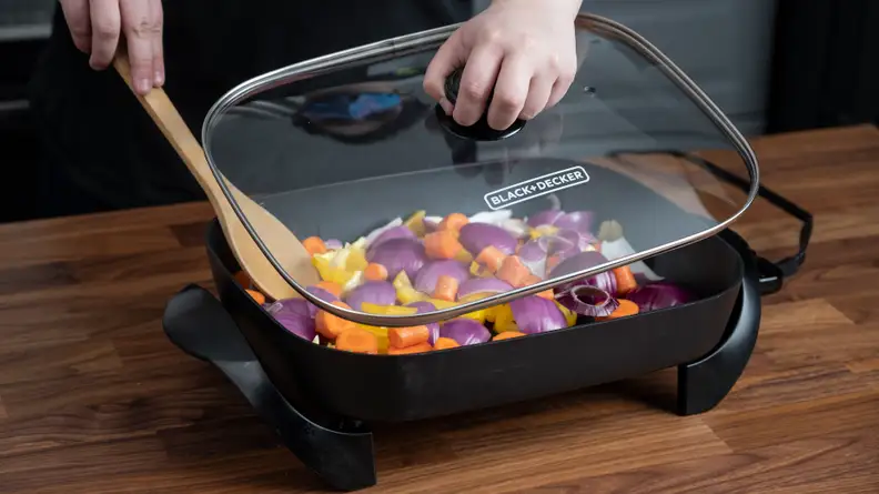 Person removing the lid from Black and Decker electric skillet to stir vegetables with wooden spoon