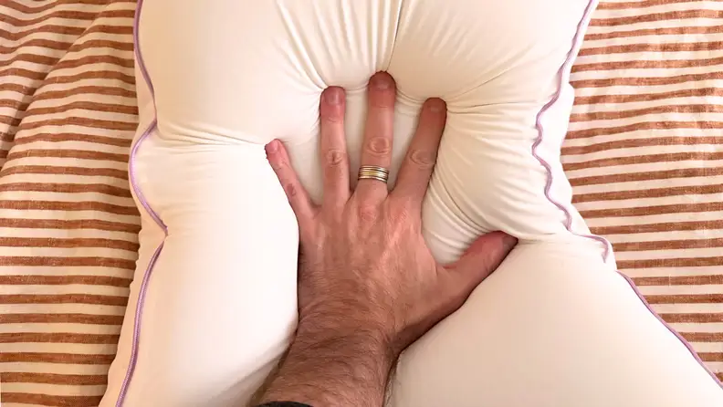 Man's hand pushing down on the center of the Layla BioCrystal Side Sleeper Pillow on top of red and tan striped sheets.