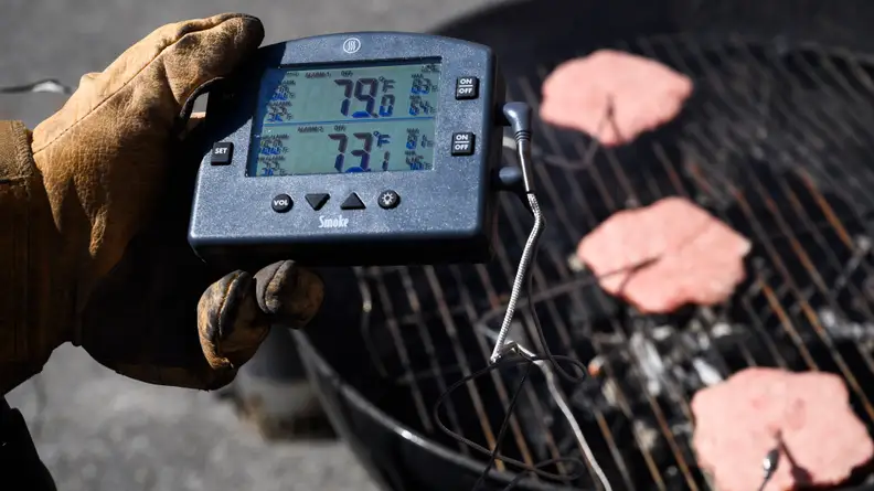 Someone holding a thermometer to check the temperature of three burgers on a grill