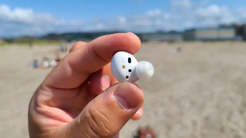 Samsung Galaxy Buds 2 is held with the interior showing against a sunny beach and mountains.