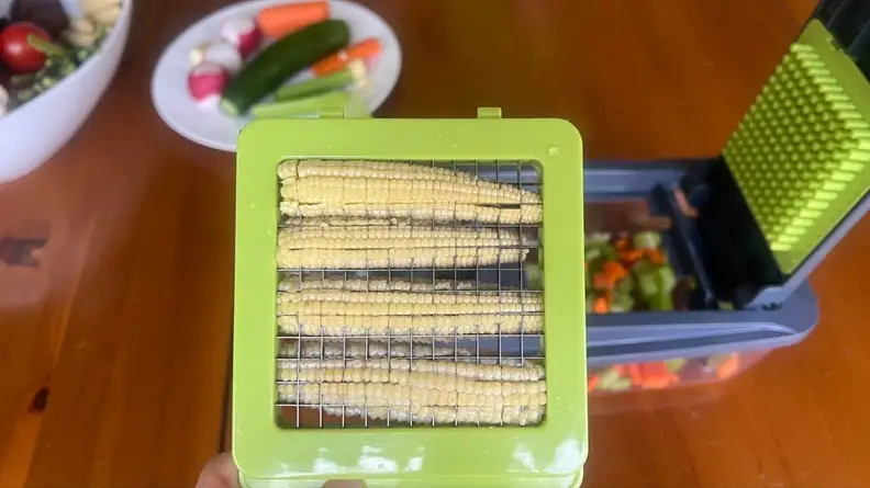 Four ears of baby corn stuck in the slicer of the Mueller vegetable chopper next to bowl and plate filled with assorted vegetables.
