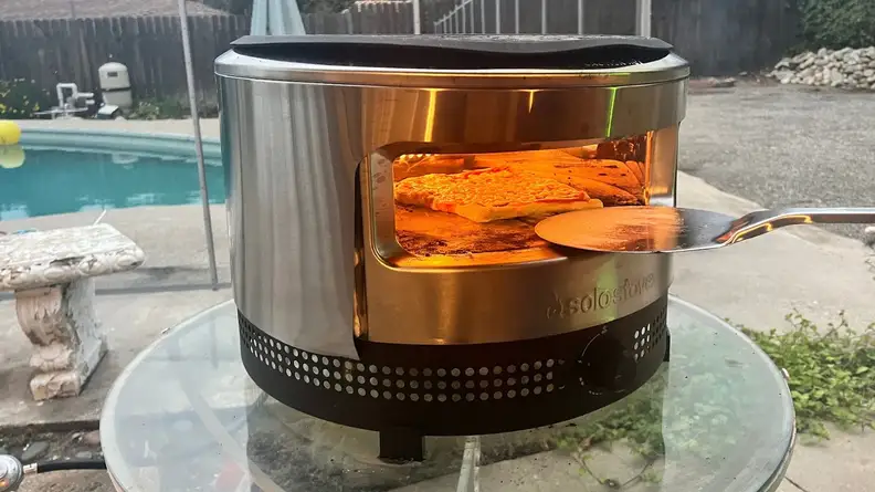 Metal pizza peel being inserted into Solo Pi Prime where pizza is cooking inside.