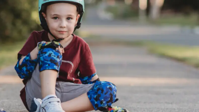A boy sits on the ground wearing blue elbow and knee pads