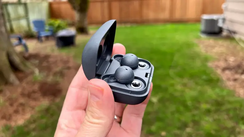 The dark gray Linkbuds sit in an open case, their double-looped design showing silver rings at the bottom in front of a green backyard.