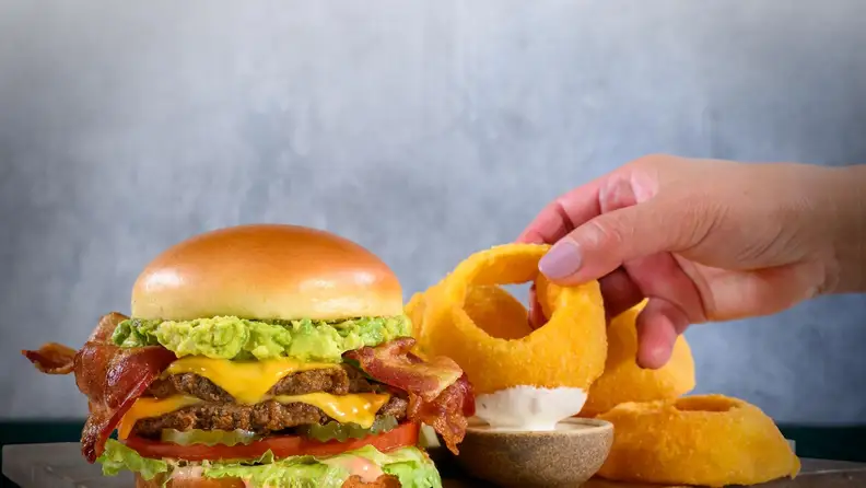 A hand reaching for onion rings next to a bacon burger with guacamole