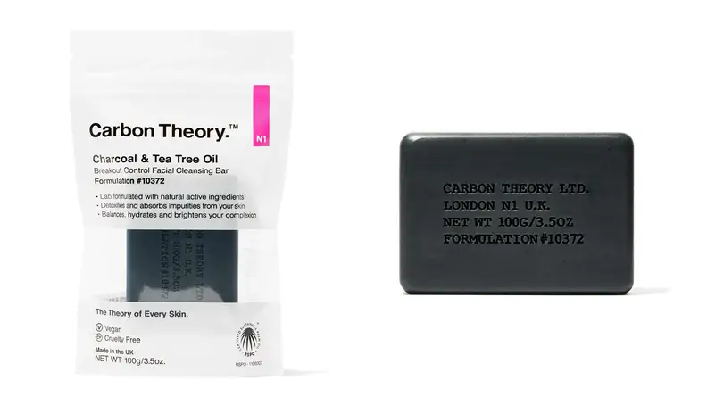 On the left: The Carbon Theory Charcoal & Tea Tree Oil Break-Out Control Facial Cleansing Bar inside its packaging sits on a white background. On the right: The dark gray Carbon Theory Charcoal & Tea Tree Oil Break-Out Control Facial Cleansing Bar sits on a white background. it has words imprinted on to it.