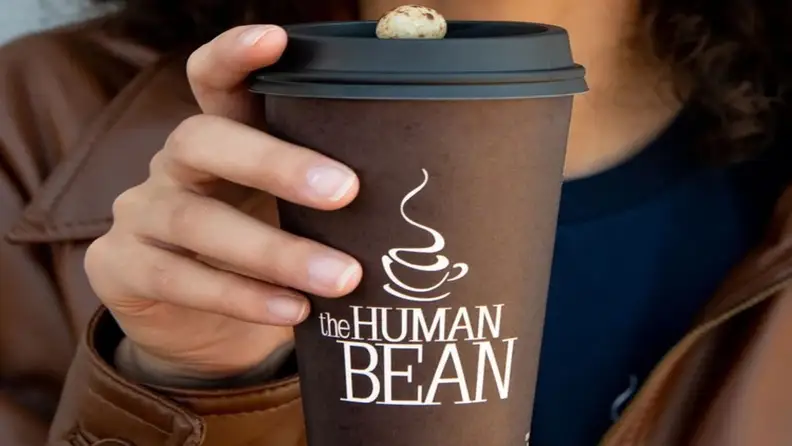 Person holding a brown Human Bean coffee cup with a lid