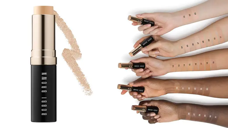 On the left, the Bobbi Brown Skin Foundation Stick stands in a tube without a cap to expose a pale-colored cream foundation and a swatch of the same color behind it. On the right, five arms ranging from fair to deep have swatches of the shades on them. Each hand holds a foundation stick.