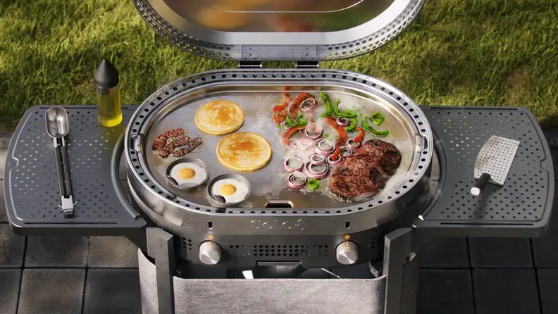 Outdoor flat-top griddle cooking pancakes, eggs, steak, and vegetables with the lid open.