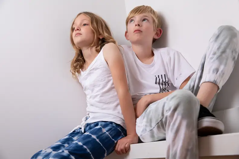 Two young children sit on a ledge wearing white shirts and tie dye pants