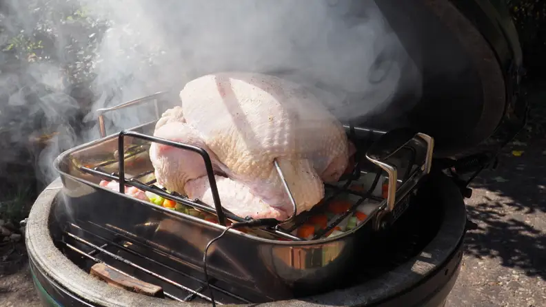 A whole raw turkey sits in a roasting pan on the grate of a kamado grill, primed for smoking.