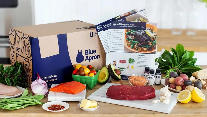 A Blue Apron box with surrounding ingredients, including produce and fresh fish, sitting on a countertop